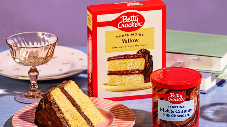 A box of Betty Crocker Super Moist Yellow cake mix sits beside a can of frosting, a slice of prepared cake, serving dishes, and cookbooks