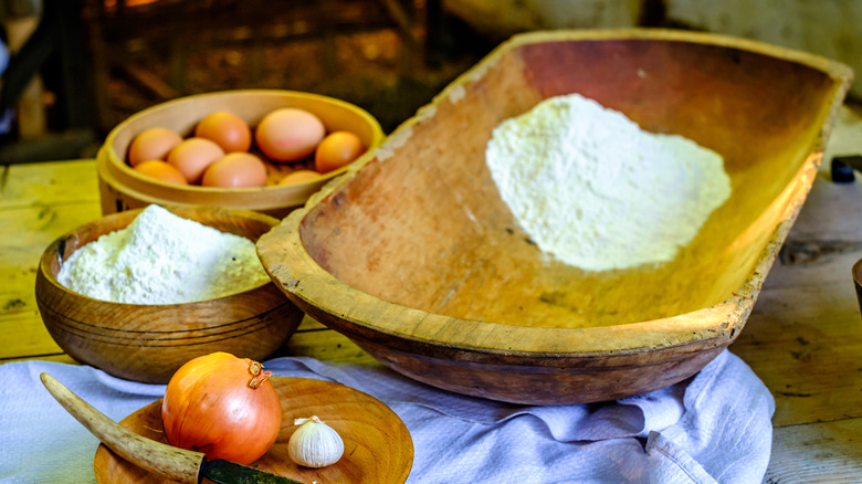 Old wooden dough bowl next to eggs and flour