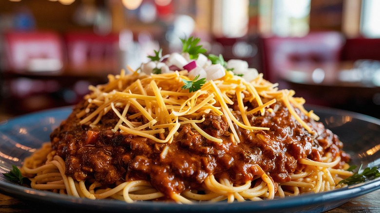 Chili ladled over a bowl of spaghetti piled with shredded cheese and diced onions