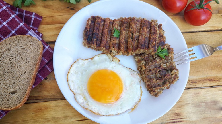 Two pieces of Goetta served with an egg over easy near a piece of toast and tomatoes on the vine