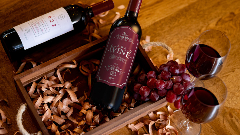 Two bottles of red wine sit in a wooden box next to red grapes and two glasses of wine