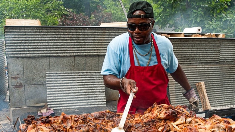 Pitmaster Rodney Scott grilling whole hog BBQ outdoors with red apron, sunglasses, and backwards hat on