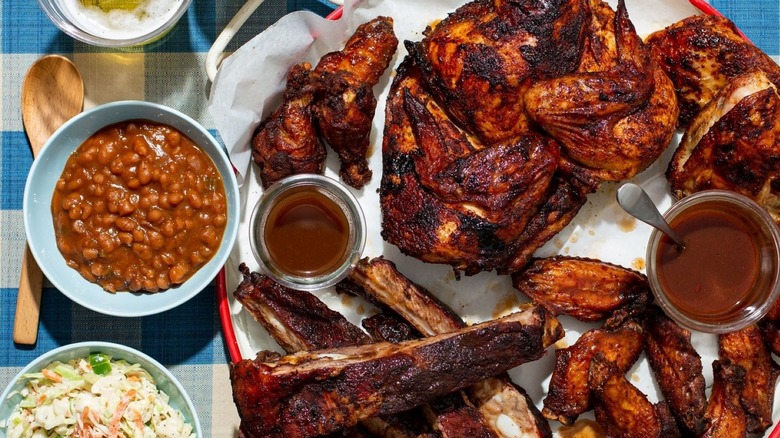 Platter of BBQ chicken, ribs, and wings, with beans on the side, on a blue plaid tablecloth