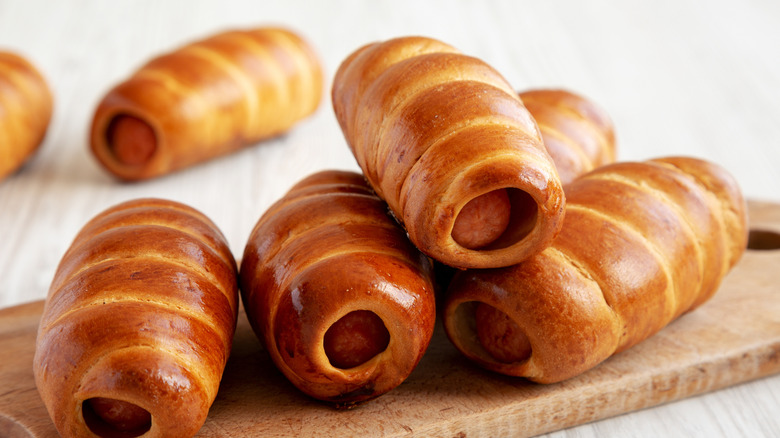 Several dough-covered sausages sit on a wooden cutting board on a white surface.