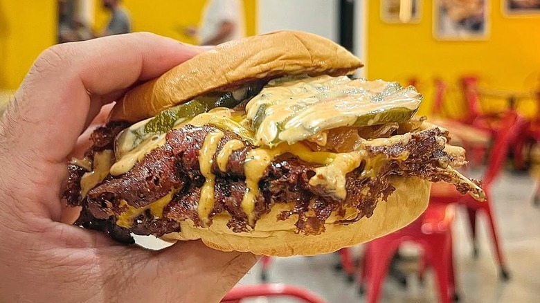 Hand holding an Easy Street burger with a thin patty, orange-yellow sauce, and pickles on a bun in front of the Easy Street sign