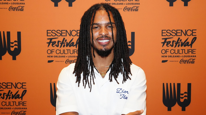 Keith Lee wearing a white short-sleeve top in front of an orange Essential Festival promotional background