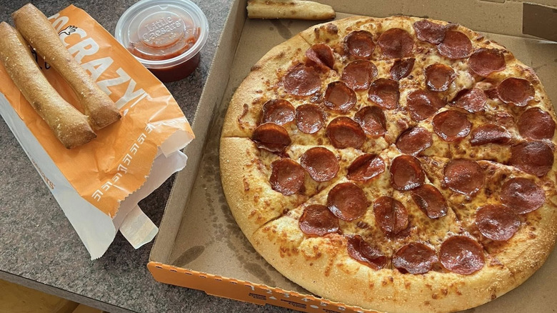 A Little Caesar's pepperoni pizza sits next to breadsticks and dip.