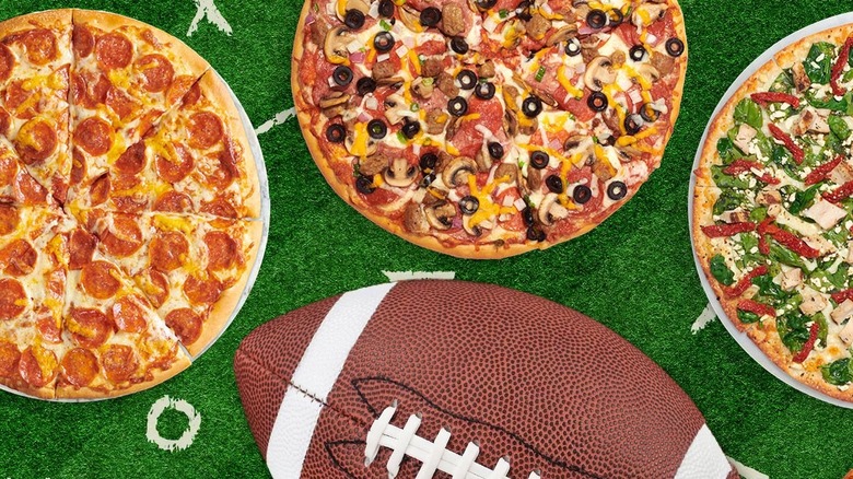 Three pizzas (pepperoni, everything lovers, and caesar salad) sit on a football field next to a football.