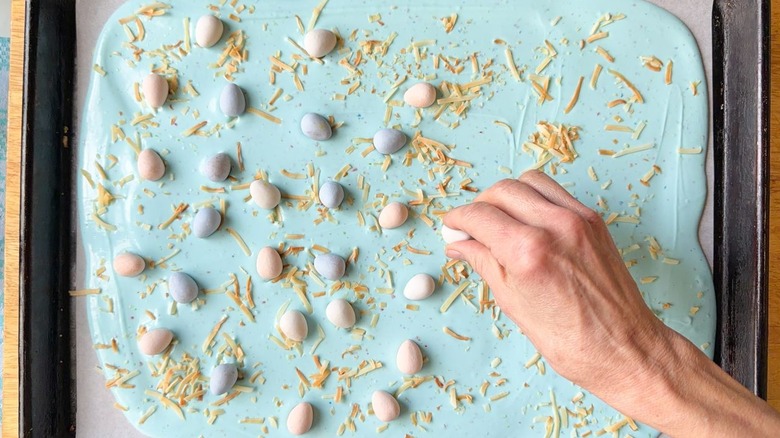 Adding candy-coated chocolate eggs to pastel blue Easter bark on sheet pan