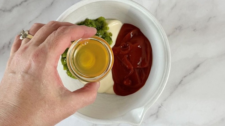 Hand holding small bowl of apple cider vinegar over bowl with relish, mayonnaise, and ketchup