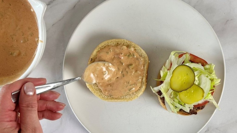 Spoon spreading burger sauce onto top bun with burger ingredients on other bun