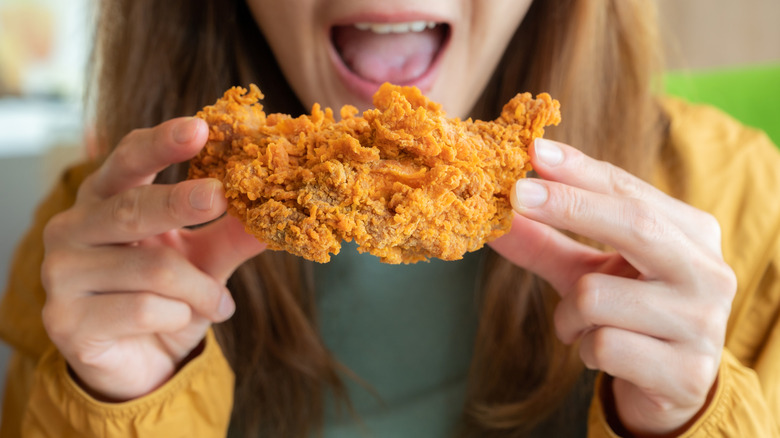 A women holds a piece of fried chicken between both hands, poised midair near her open mouth to take a bite