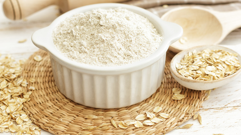 A bowl of oat flour sitting on a straw mat with a spoon of oats to the right
