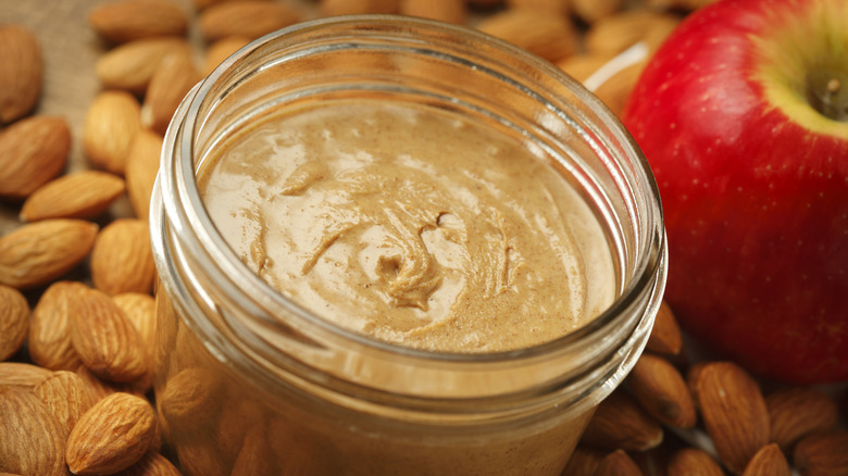A jar of almond butter next to loose almonds and an apple.