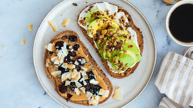 Two slices of toast, one with avocado and seeds, and the other with peanut butter, dried fruit, and coconut