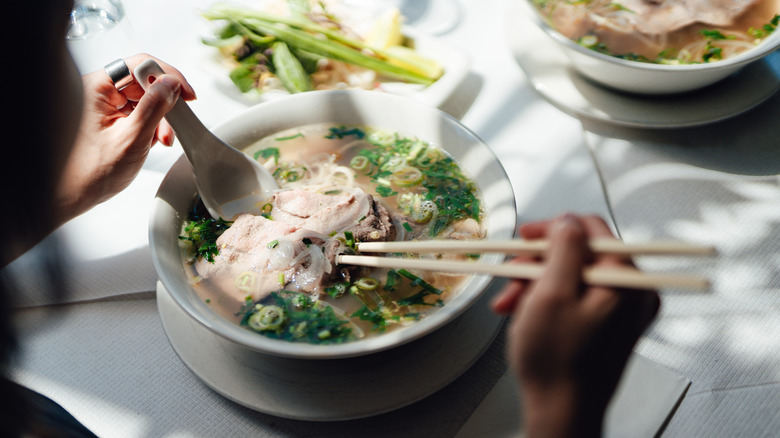 Over the shoulder shot of a person holding chopsticks and a spoon in a bowl of pho