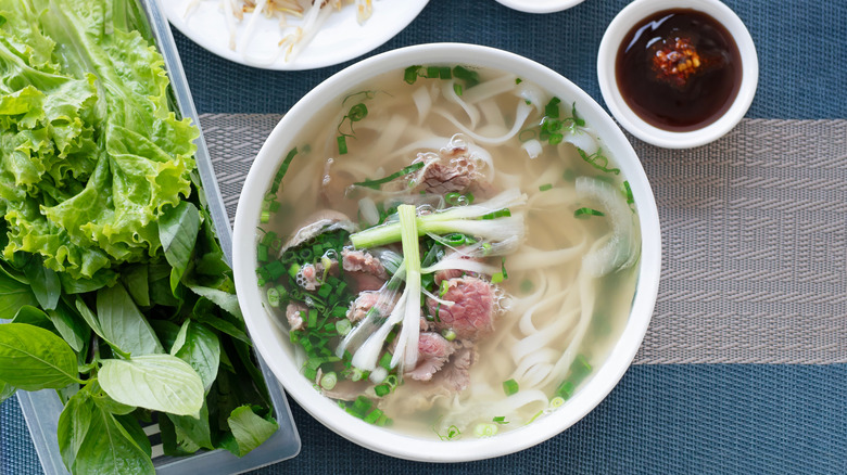 A bowl of beef pho sits next to a plate of greens and a small bowl of chili sauce