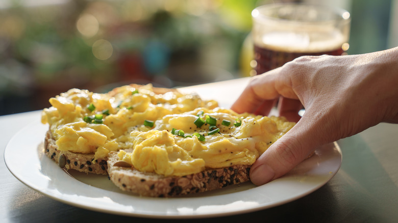 person's hand is reaching for toast topped with scrambled eggs that's on a white plate
