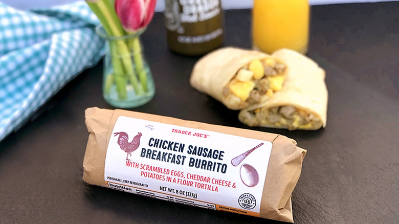 A Trader Joe's Chicken Sausage Breakfast Burrito package with a prepared burrito, a vase of flowers, and a checkered napkin in the background