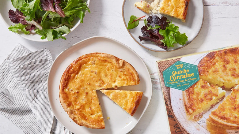 A Quiche Lorraine from Trader Joe's sits on a white plate next to the product package, a plate of salad, and another plate containing salad greens and a wedge of quiche