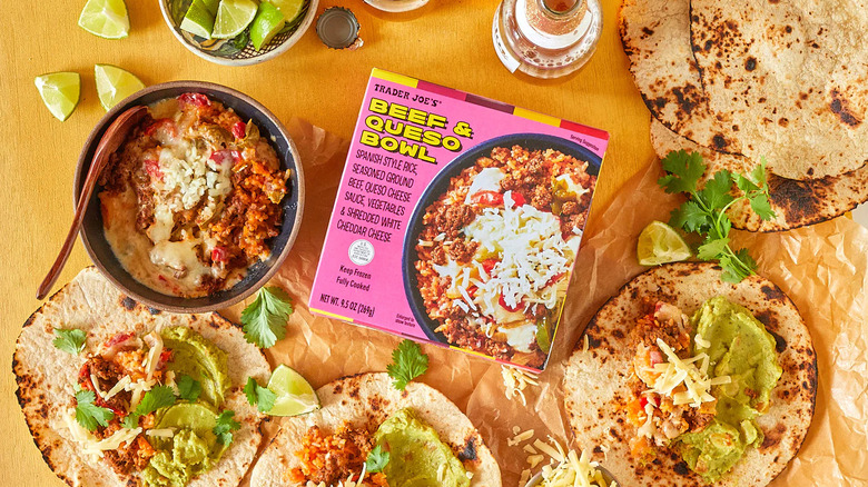 A Trader Joe's Beef & Queso Bowl package is surrounded by a prepared bowl, tortillas, lime wedges, cilantro, and other accoutrements
