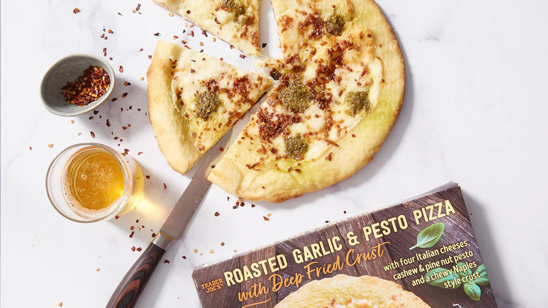 A prepared Trader Joe's Roasted Garlic & Pesto Pizza with Deep Fried Crust sits near a product box, a knife, and ramekins of oil and pepper flakes