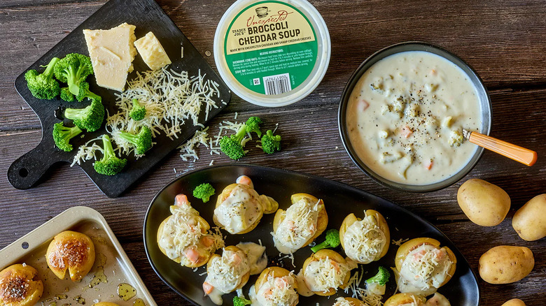 A container of Trader Joe's Unexpected Broccoli Cheddar Soup sits next to a bowl of soup, a cutting board with broccoli and cheese, raw potatoes, and pans of cooked potatoes