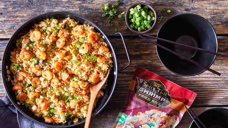 A pan of Trader Joe's Spicy Thai Shrimp Fried Rice sits beside an empty bowl with chopsticks, a ramekin of scallions, and the product package