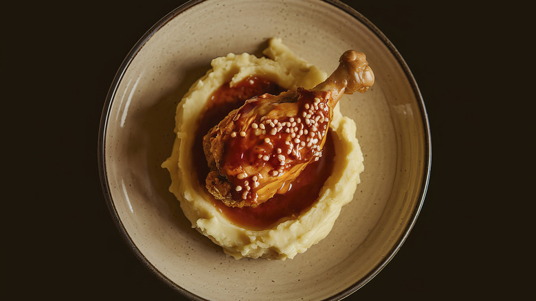 A plate is topped with mashed potatoes, a chicken drumstick, chicken gravy, and what could be sesame seeds