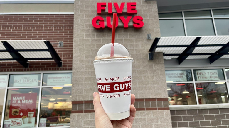 Hand holding Five Guys Chocolate Milkshake in front of storefront