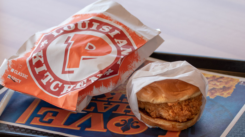 A Popeye's branded food bag sits next to a wrapped fried chicken sandwich on a fast food tray