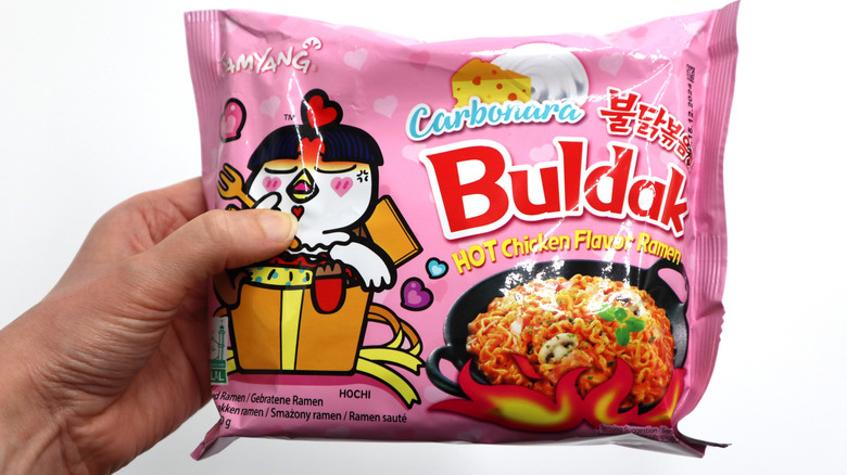 A person holds a pink Buldak ramen carbonara package.