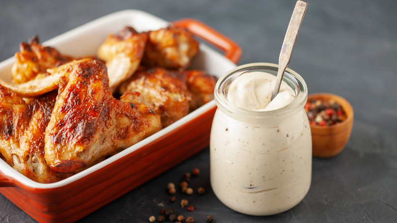 A small bottle of Alabama white sauce with a spoon sits next to a casserole dish filled with grilled chicken wings