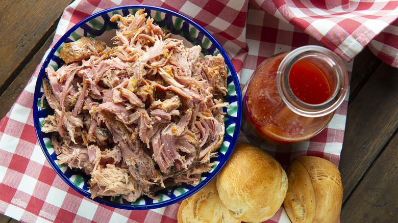 A bowl of pulled pork sits next to a bottle of Eastern Carolina barbecue sauce and rolls