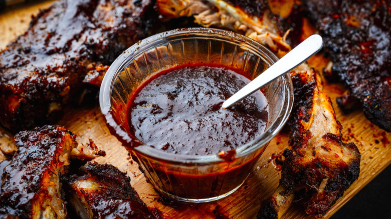 A bowl of Kansas City barbecue sauce with a spoon sits on a wooden board surrounded by charred ribs
