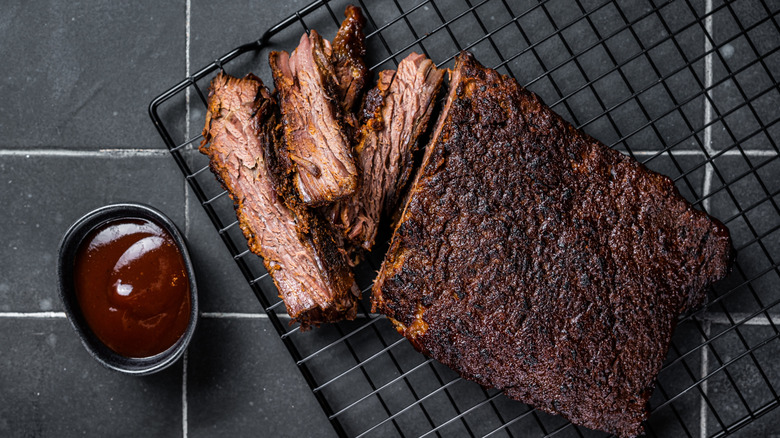 A smoked brisket on a cooling rack rests next to a small bowl of Texas barbecue sauce