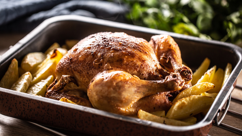 A roast chicken in a roasting dish on top of sliced potoatoes