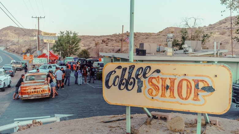 A sign saying coffee shop, in the desert, with lots of people outside and vintage rusty cars