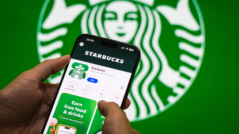 Starbucks app download screen on iPhone in front of larger LED Starbucks logo display.
