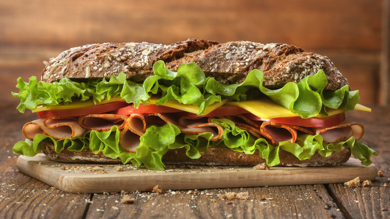 An enormous sandwich filled with meat, cheese, and lettuce sits on a wooden cutting board
