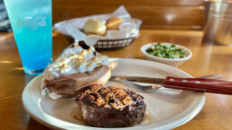 A filet mignon steak sits on a plate with a roasted sweet potato and a side of green beans and bread, with a neon blue drink at the table