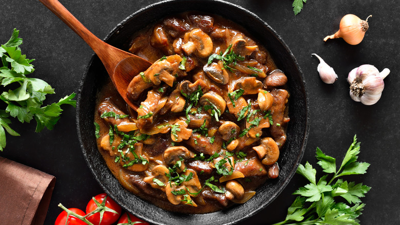 Beef stroganoff in a black skillet surrounded by herbs, aromatics, and tomatoes.