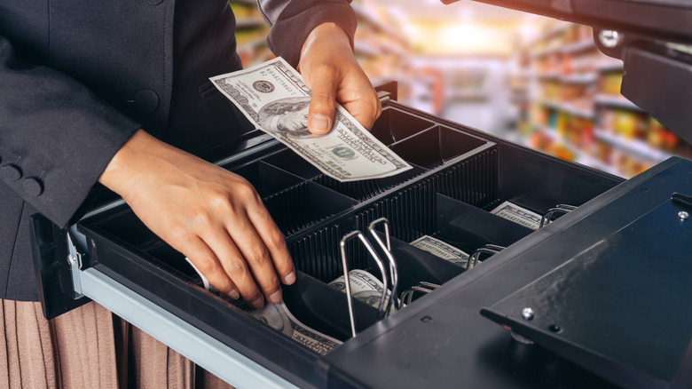 A person places a $100 bill into an open cash drawer