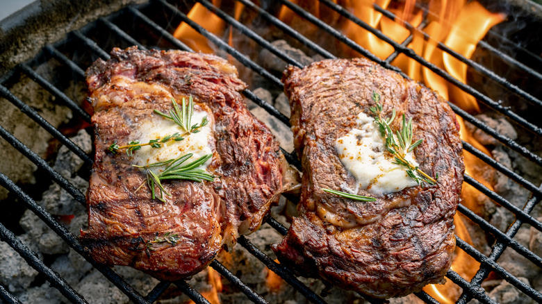 Two ribeye steaks cooking on a grill with butter and rosemary.