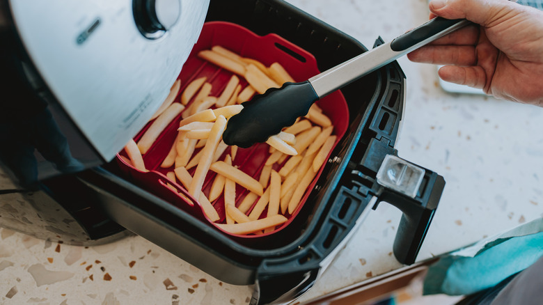 A hand holding a pair of tongs agitates fries in a silicone air fryer liner