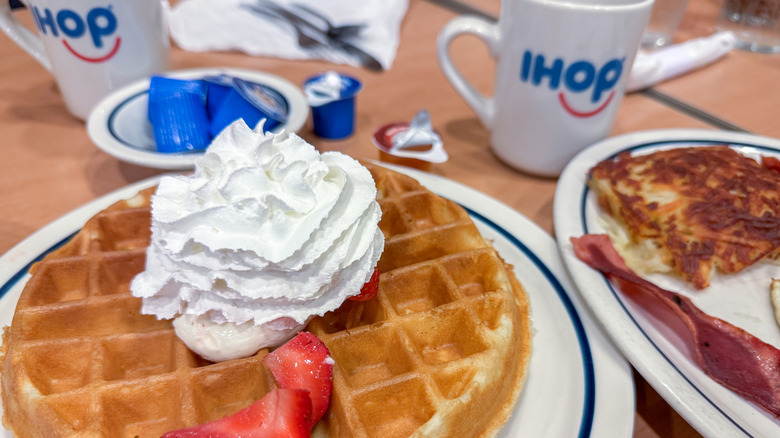 Waffle topped with strawberry and whip cream on an IHOP table with mugs and another plate