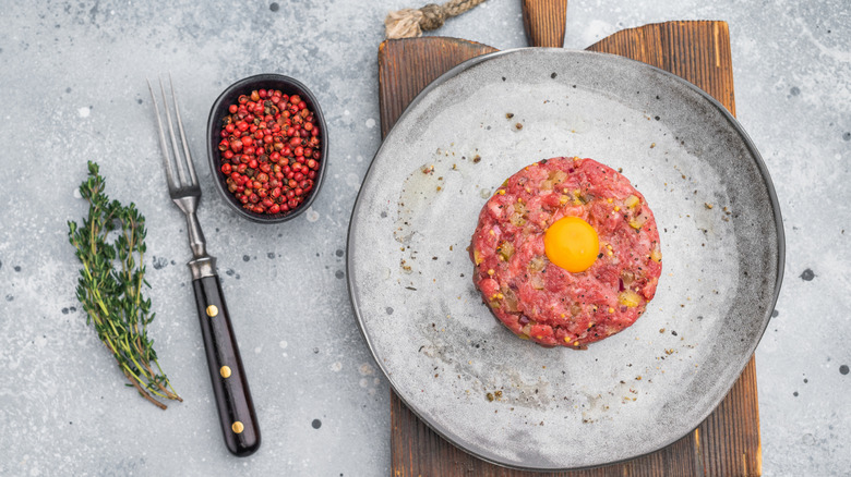 Chopped beef with an egg yolk sits on a plate next to fresh herbs and pink peppercorns.
