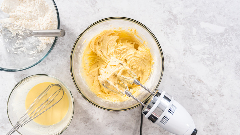 A batch of yellow cake mix sits in a glass bowl next to a bowl of flour and a bowl of beaten eggs
