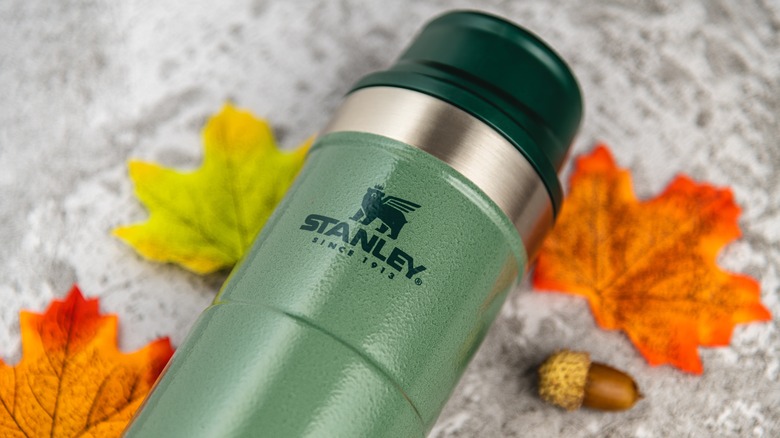 Close up on a green Stanley thermos with a trigger action lid  surrounded by orange and yellow leaves