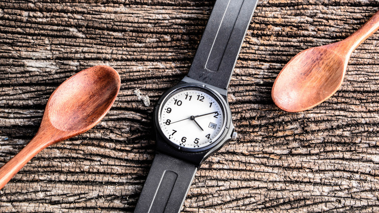 An analog wristwatch lies between two wooden spoons on a wooden board
